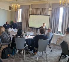 Group of people sat at a table watching a trainer presenting in front of a large screen
