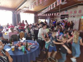 A busy community hall filled with adults and children gathered around round tables decorated with Union Jack flags and bunting for a celebration event. Guests of all ages are eating, chatting, and dancing, with children playing energetically in the foreground. A "Friends of Families" promotional banner is visible in the background.
