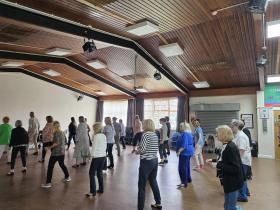 A large group of adults taking part in a line dancing class inside the main hall at Abram Community Centre. Participants of various ages are spread across the wooden floor in informal rows. An Abram Community Centre banner is partially visible on the wall in the background.