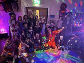 A group of children and adults posing together at a Halloween party inside a community hall, lit up with colourful disco lights. The children are dressed in a variety of Halloween costumes and are smiling and giving thumbs up to the camera. The room is decorated with spooky props and atmospheric lighting.
