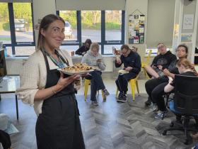 A lady is holding a plate with pastries. A group of 5 students enjoy the pastries in the classroom while having a break. 