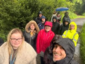 A group of 12 young people pose for a selfie taken by an adult man outdoors, during a hike. Most of them wear wet raincoats and are surrounded by bushes.