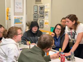 The picture shows a classroom. A group of young people are gathered around a table holding markers and pens, awaiting for the supervisor's instructions.