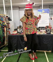 A black woman posing with custom tailored Christmas hat, top, skirt, with more colourful dresses and outfits on display in the background