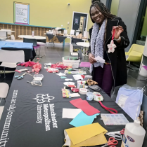 A woman stands behind a craft table at a community workshop, holding up a handmade fabric ornament. The table is covered with sewing and crafting materials, including fabric pieces, scissors, ribbons, and tools, demonstrating sustainable fashion and creative skills activities at Manchester Metropolitan University.