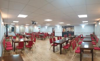 A spacious, well-lit function room set up with rows of dark wooden tables and red chairs on a wooden floor, with a bar area visible at the far end. The room appears ready to host a large event or gathering.