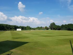 A well-maintained cricket ground with neatly mown grass under a bright blue summer sky with white clouds, surrounded by trees. Batting screens, practice nets, and club facilities are visible in the background.