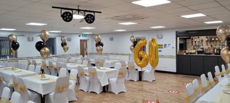 The organisation's function room decorated for a 60th birthday celebration, with white chair covers tied with gold ribbon, balloon centrepieces in black and gold, and large gold "60" number balloons in the centre of the room.