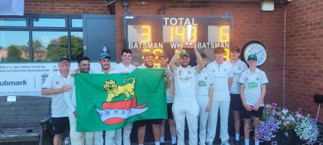 A cricket team in white kit celebrating a victory outside the clubhouse, with one player holding a trophy aloft and others displaying a green flag bearing a lion crest, in front of an illuminated scoreboard.