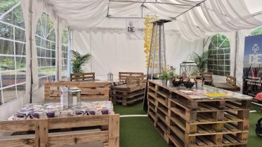 The interior of a decorated marquee set up as a relaxed event space, with furniture made from wooden pallets, cushioned seating, potted plants, and lanterns on artificial grass, with a Duke of Edinburgh award banner visible in the background.