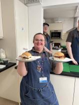 A smiling young trainee wearing a denim apron and the organisation's lanyard proudly holds up two plates of bacon rolls in a café kitchen, with two colleagues visible working behind her.
