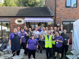 A large group of staff, trainees, and volunteers giving thumbs up outside the entrance to the Nest Café on a sunny day, with the café's lilac awning and signage visible above the door.