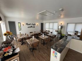 The bright, modern interior of the Nest Café, with wooden tables and brown chairs arranged across a wood-effect floor, warm wall lighting, sunflowers on the counter, and the café's name displayed in decorative lettering on the wall.