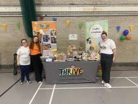 Three ladies stand by a table that displays Thrive CIC's items and accessories for sale. The background shows two banners promoting the organisation. The venue has been decorated with balloons and bunting.