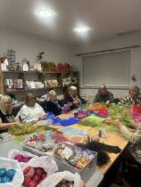 A group of seven middle-aged ladies is sat around a big table. They are making handmade ornaments with colourful fabrics placed in the middle of the table. There are other materials like Christmas baubles and other Christmassy elements on the side. 