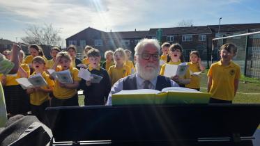 A musician plays a piano outdoors while a large group of primary school children in yellow uniforms sing from sheet music behind him in a sunny school playground.