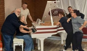 A group of six adults laughing and gathered around a striking white grand piano decorated with Wigan Warriors rugby club branding, during what appears to be a rehearsal or informal session.