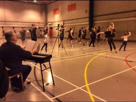 A pianist plays a keyboard at the side of a sports hall while a large group of dancers in black rehearse an energetic routine across the floor, with a camera on a tripod recording the session.