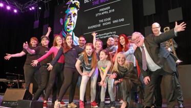 A lively group of performers of various ages pose excitedly on a theatre stage in front of a large screen promoting a musical premiere at The Edge in Wigan, with arms outstretched and big smiles for the camera.