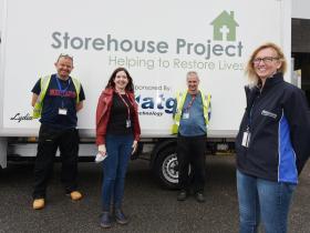 Four people smiling in front of the organisation's white delivery van, which displays the tagline "Helping to restore lives" and a sponsor's logo, in what appears to be a warehouse or distribution yard.