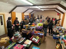 A room filled with a large collection of donated toys and games, with a group of young volunteers and adults sorting through them, appearing to be preparing donations for distribution.