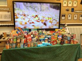 A table in a primary school classroom piled high with a wide variety of donated tinned and packaged food items, collected as part of a food drive, with a large interactive screen and classroom displays visible in the background.
